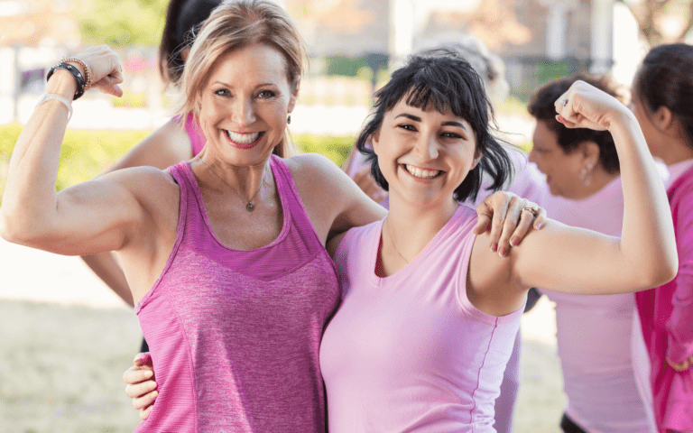 Two women flexing their muscles, wearing pink and happily smiling while looking at the camera.