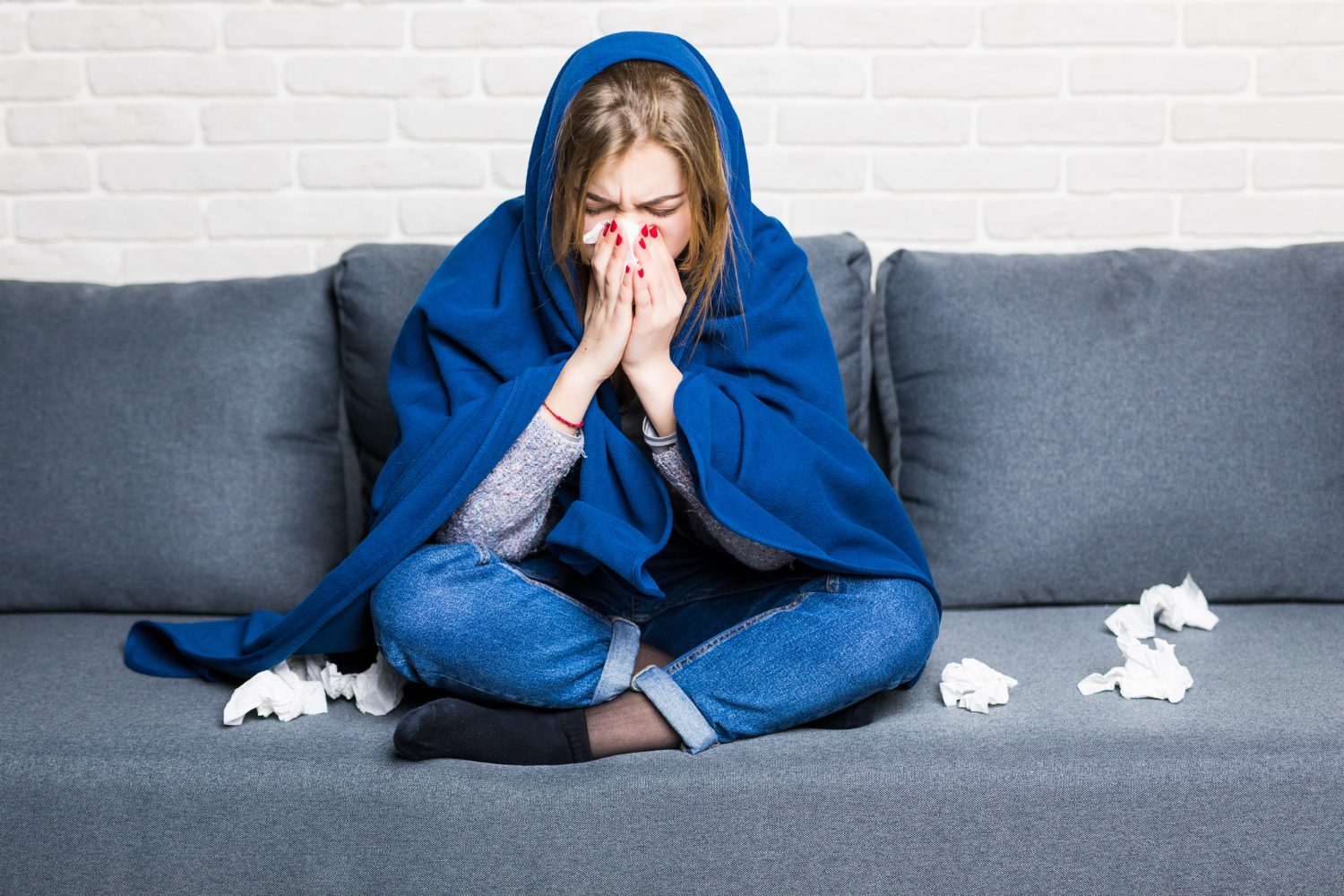 Sick woman covered herself in blue blanket sitting in her couch holding a napkin to cover her nose.